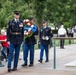 A military wreath-laying ceremony is taking place at what appears to be a national memorial or cemetery, likely Arlington National Cemetery. Uniformed U.S. Army soldiers in dress blues are carrying a red, white, and blue floral wreath while civilians and veterans observe from the sides. The formal procession suggests a commemorative or memorial event.