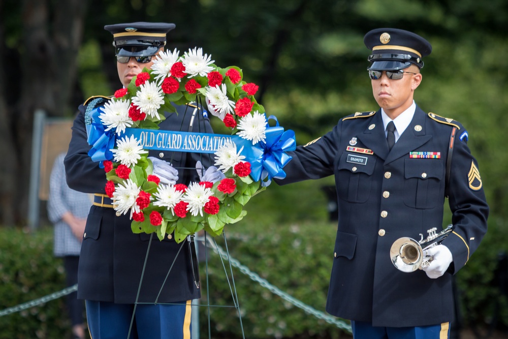 Two U.S. Army soldiers in dress uniforms are participating in a formal wreath-laying ceremony. One soldier holds a floral wreath bearing a blue ribbon reading 'Old Guard Association,' while the other holds a bugle, suggesting a ceremonial musical performance such as 'Taps.' The setting appears to be an outdoor memorial or military ceremony.