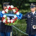 Two U.S. Army soldiers in dress uniforms are participating in a formal wreath-laying ceremony. One soldier holds a floral wreath bearing a blue ribbon reading 'Old Guard Association,' while the other holds a bugle, suggesting a ceremonial musical performance such as 'Taps.' The setting appears to be an outdoor memorial or military ceremony.