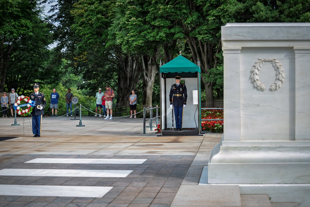 This image depicts a ceremonial event at the Tomb of the Unknown Soldier at Arlington National Cemetery, Virginia. A uniformed soldier stands guard in the sentry box while another soldier holds a memorial wreath nearby. Civilian visitors observe the solemn ceremony from behind a rope barrier in the background.