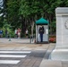 This image depicts a ceremonial event at the Tomb of the Unknown Soldier at Arlington National Cemetery, Virginia. A uniformed soldier stands guard in the sentry box while another soldier holds a memorial wreath nearby. Civilian visitors observe the solemn ceremony from behind a rope barrier in the background.