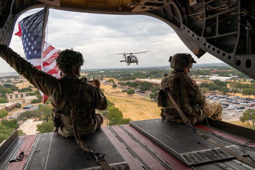 Two U.S. Army soldiers in full combat gear sit on the open rear ramp of a large military helicopter, likely a CH-47 Chinook, in flight over what appears to be a military installation or base. One soldier is holding an American flag that is extended outward in the wind. A second helicopter, identifiable as a UH-60 Black Hawk, is visible flying in formation nearby.