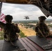 Two U.S. Army soldiers in full combat gear sit on the open rear ramp of a large military helicopter, likely a CH-47 Chinook, in flight over what appears to be a military installation or base. One soldier is holding an American flag that is extended outward in the wind. A second helicopter, identifiable as a UH-60 Black Hawk, is visible flying in formation nearby.