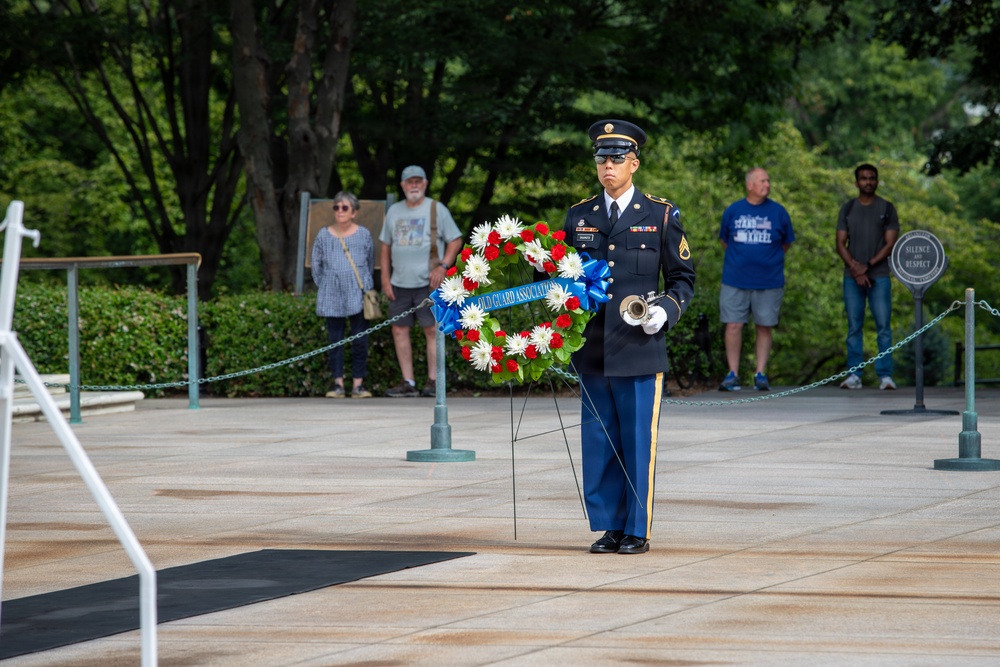 A U.S. Army soldier in dress blues stands at attention holding a bugle in front of a ceremonial wreath at what appears to be Arlington National Cemetery. The wreath bears a blue ribbon reading 'Old Guard Association,' indicating a formal military memorial ceremony. Civilian onlookers stand behind a rope barrier observing the solemn event.