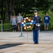 A U.S. Army soldier in dress blues stands at attention holding a bugle in front of a ceremonial wreath at what appears to be Arlington National Cemetery. The wreath bears a blue ribbon reading 'Old Guard Association,' indicating a formal military memorial ceremony. Civilian onlookers stand behind a rope barrier observing the solemn event.
