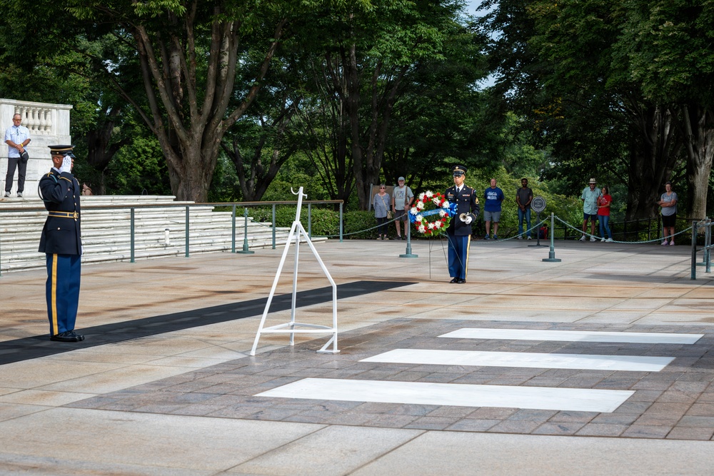 A formal military wreath-laying ceremony is taking place at what appears to be the Tomb of the Unknown Soldier at Arlington National Cemetery. Two uniformed soldiers are participating in the ceremony — one standing at attention as a sentinel and one carrying a floral wreath — while civilian spectators observe from behind a rope barrier. The setting features a large stone plaza with white painted markers, marble structures, and lush green trees in the background.