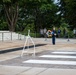 A formal military wreath-laying ceremony is taking place at what appears to be the Tomb of the Unknown Soldier at Arlington National Cemetery. Two uniformed soldiers are participating in the ceremony — one standing at attention as a sentinel and one carrying a floral wreath — while civilian spectators observe from behind a rope barrier. The setting features a large stone plaza with white painted markers, marble structures, and lush green trees in the background.
