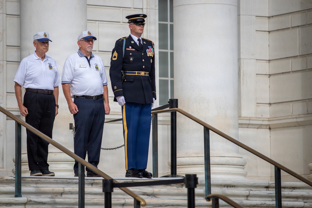 Three men stand on the steps of a large neoclassical building with white marble columns. A uniformed U.S. Army soldier in formal dress blues stands at attention alongside two older civilians wearing matching white polo shirts with insignia patches and baseball caps. The setting and attire suggest a formal military ceremony or memorial event.