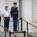 Three men stand on the steps of a large neoclassical building with white marble columns. A uniformed U.S. Army soldier in formal dress blues stands at attention alongside two older civilians wearing matching white polo shirts with insignia patches and baseball caps. The setting and attire suggest a formal military ceremony or memorial event.