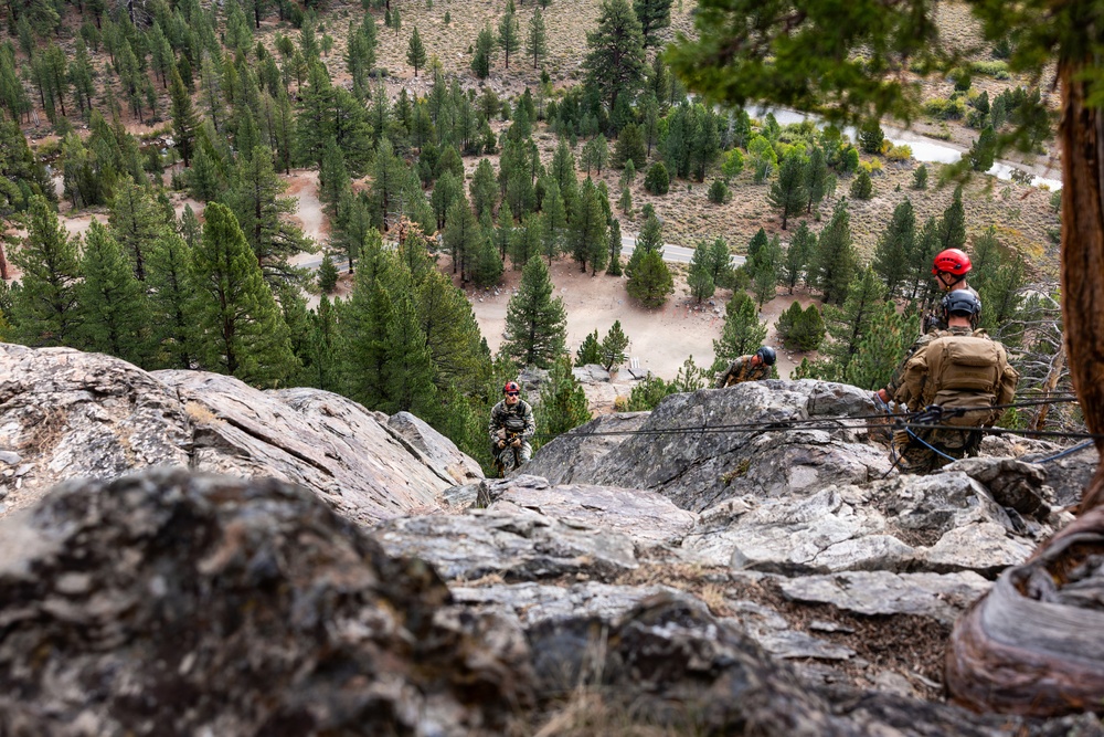 Military personnel in camouflage uniforms and tactical gear are conducting a rappelling or rock climbing exercise on a steep rocky cliff face. The scene appears to be a training exercise in a mountainous, forested environment with conifer trees visible in the valley below. At least three individuals are visible at various positions on the rock face, equipped with ropes, helmets, and body armor.