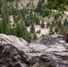 Military personnel in camouflage uniforms and tactical gear are conducting a rappelling or rock climbing exercise on a steep rocky cliff face. The scene appears to be a training exercise in a mountainous, forested environment with conifer trees visible in the valley below. At least three individuals are visible at various positions on the rock face, equipped with ropes, helmets, and body armor.