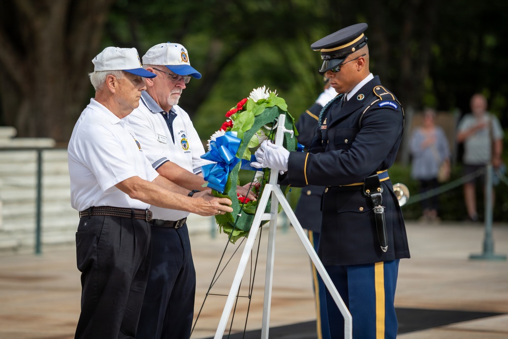 A formal wreath-laying ceremony is taking place, likely at Arlington National Cemetery or a similar military memorial site. Two civilians wearing matching white polo shirts and blue-brimmed caps are presenting a floral wreath to a U.S. Army Honor Guard soldier in dress uniform. The wreath features red, white, and green flowers with a prominent blue ribbon bow.