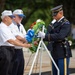 A formal wreath-laying ceremony is taking place, likely at Arlington National Cemetery or a similar military memorial site. Two civilians wearing matching white polo shirts and blue-brimmed caps are presenting a floral wreath to a U.S. Army Honor Guard soldier in dress uniform. The wreath features red, white, and green flowers with a prominent blue ribbon bow.