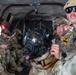 Military personnel are seated inside what appears to be a military helicopter or transport aircraft. A military working dog (MWD), likely a Belgian Malinois, is wearing specialized protective gear including a canine tactical helmet with eye protection and a muzzle. The soldiers are wearing MultiCam (OCP) uniforms with unit patches visible, including what appears to be a 1st Cavalry Division patch.