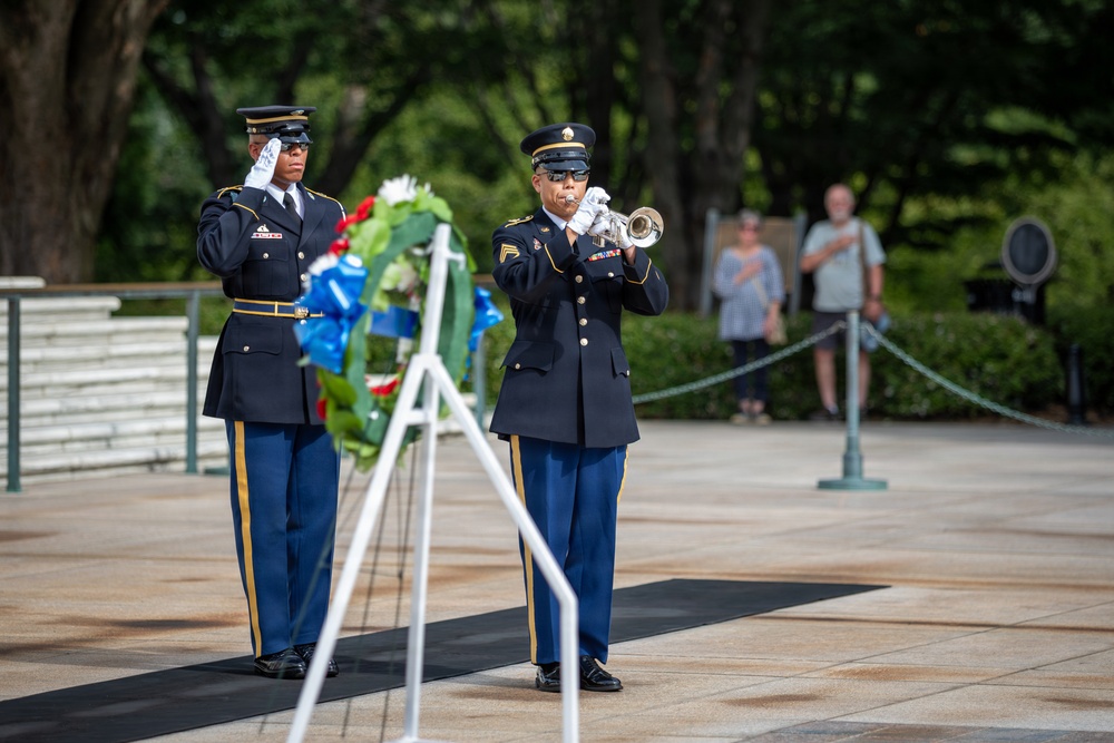 A formal military ceremony is taking place at what appears to be a memorial or monument, likely Arlington National Cemetery or a similar venue. Two U.S. Army soldiers in dress blue uniforms are performing official duties — one playing a bugle (likely 'Taps') and one rendering a salute beside a floral wreath on a stand. Civilian observers stand respectfully in the background, one with hand over heart.