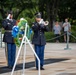 A formal military ceremony is taking place at what appears to be a memorial or monument, likely Arlington National Cemetery or a similar venue. Two U.S. Army soldiers in dress blue uniforms are performing official duties — one playing a bugle (likely 'Taps') and one rendering a salute beside a floral wreath on a stand. Civilian observers stand respectfully in the background, one with hand over heart.