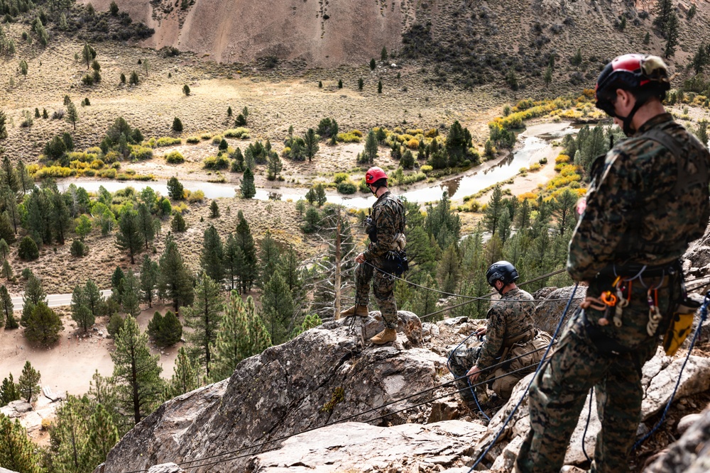 Three individuals in military camouflage uniforms are conducting rappelling or rope management training on a rocky cliff edge at high elevation. They are equipped with climbing helmets (red and blue) and rope systems, overlooking a scenic mountain valley with a winding river, conifer trees, and arid terrain below. The setting appears to be a military mountain warfare or climbing training exercise.
