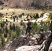 Three individuals in military camouflage uniforms are conducting rappelling or rope management training on a rocky cliff edge at high elevation. They are equipped with climbing helmets (red and blue) and rope systems, overlooking a scenic mountain valley with a winding river, conifer trees, and arid terrain below. The setting appears to be a military mountain warfare or climbing training exercise.