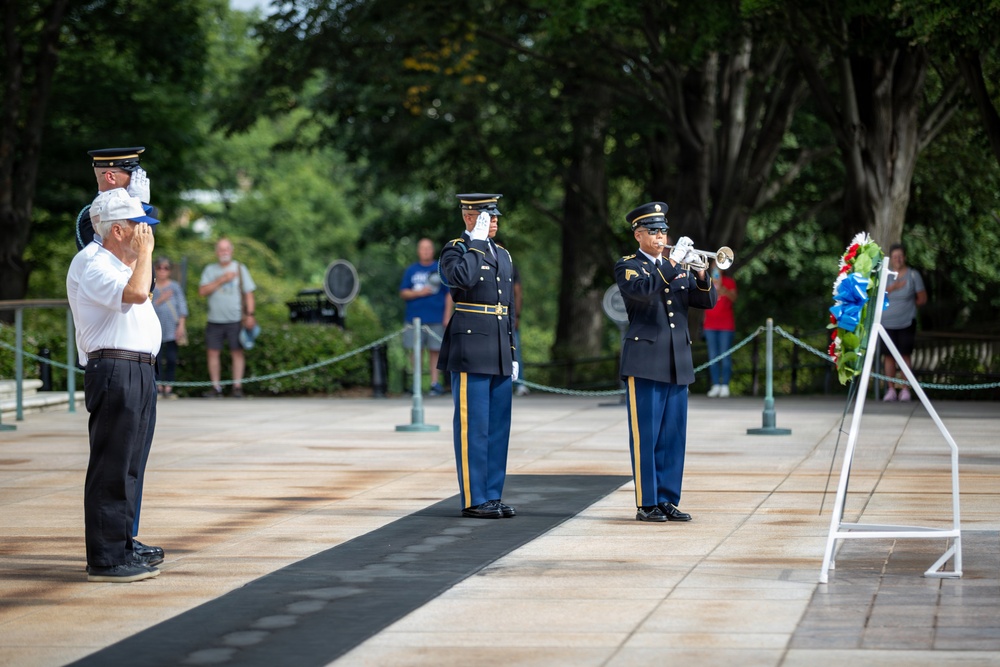A formal military wreath-laying ceremony is taking place at what appears to be the Tomb of the Unknown Soldier at Arlington National Cemetery. Two uniformed U.S. Army soldiers are saluting while a third plays a bugle, likely performing 'Taps.' A civilian veteran in a white shirt and blue cap renders a hand salute alongside the military personnel.