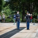 A formal military wreath-laying ceremony is taking place at what appears to be the Tomb of the Unknown Soldier at Arlington National Cemetery. Two uniformed U.S. Army soldiers are saluting while a third plays a bugle, likely performing 'Taps.' A civilian veteran in a white shirt and blue cap renders a hand salute alongside the military personnel.