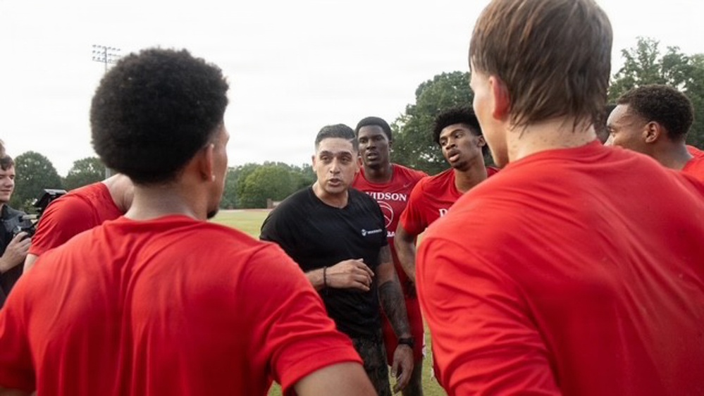 A coach or trainer dressed in a black shirt addresses a group of athletes wearing red jerseys during what appears to be an outdoor practice or training session. The group is gathered in a huddle formation on a grass field, with the coach speaking at the center. One jersey is partially visible with the text 'DAVIDSON' on it.