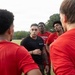 A coach or trainer dressed in a black shirt addresses a group of athletes wearing red jerseys during what appears to be an outdoor practice or training session. The group is gathered in a huddle formation on a grass field, with the coach speaking at the center. One jersey is partially visible with the text 'DAVIDSON' on it.