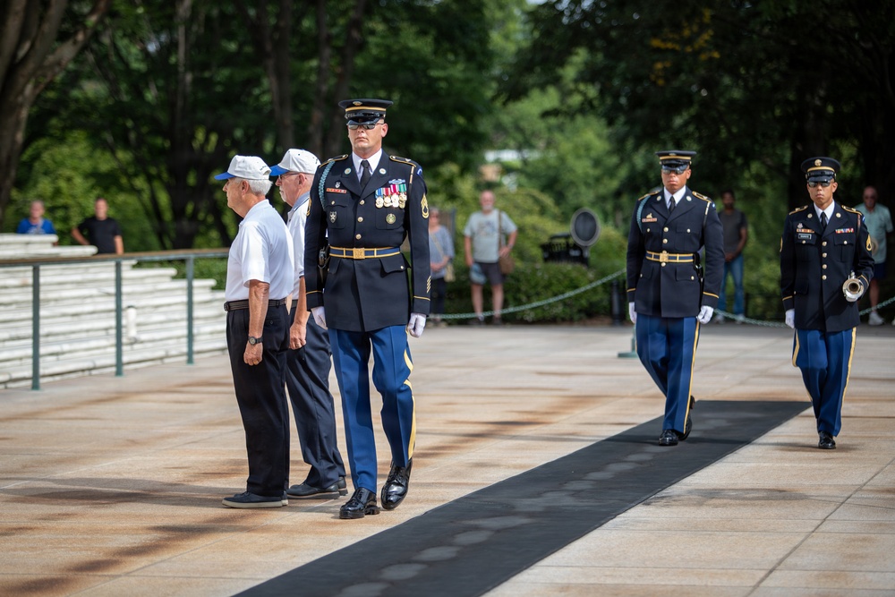 A formal military ceremony is taking place at what appears to be the Tomb of the Unknown Soldier at Arlington National Cemetery. U.S. Army soldiers in dress blue uniforms are escorting two elderly civilians, likely veterans, during a wreath-laying or memorial ceremony. A bugler is visible among the honor guard participants approaching from the right.