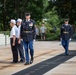 A formal military ceremony is taking place at what appears to be the Tomb of the Unknown Soldier at Arlington National Cemetery. U.S. Army soldiers in dress blue uniforms are escorting two elderly civilians, likely veterans, during a wreath-laying or memorial ceremony. A bugler is visible among the honor guard participants approaching from the right.