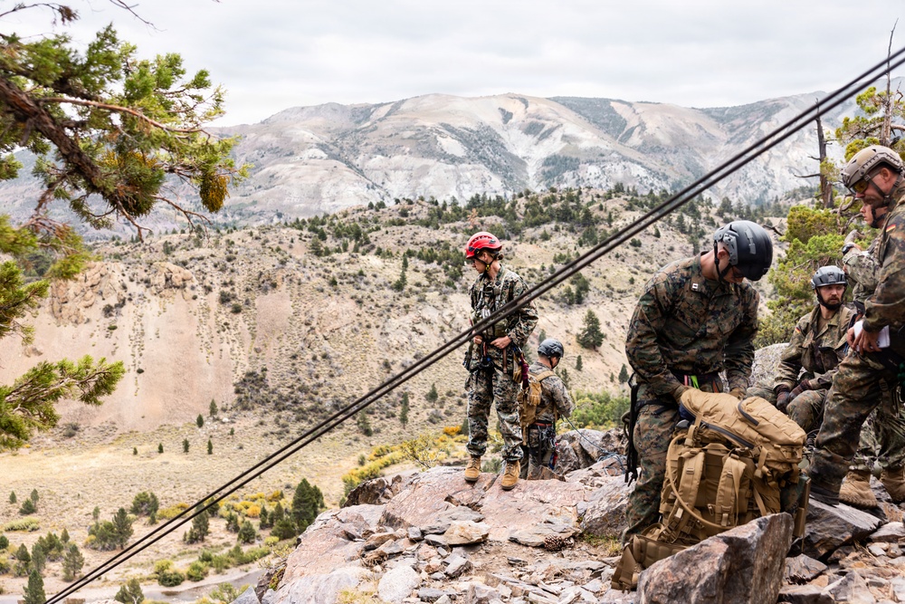 A group of military personnel in camouflage uniforms are conducting mountain climbing or rappelling training on a rocky ridge. They are equipped with helmets, harnesses, ropes, and tactical backpacks, positioned at elevation with a dramatic mountain valley visible in the background. The setting appears to be a high-altitude arid mountain environment, consistent with locations such as the Sierra Nevada or similar western U.S. ranges.
