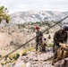A group of military personnel in camouflage uniforms are conducting mountain climbing or rappelling training on a rocky ridge. They are equipped with helmets, harnesses, ropes, and tactical backpacks, positioned at elevation with a dramatic mountain valley visible in the background. The setting appears to be a high-altitude arid mountain environment, consistent with locations such as the Sierra Nevada or similar western U.S. ranges.