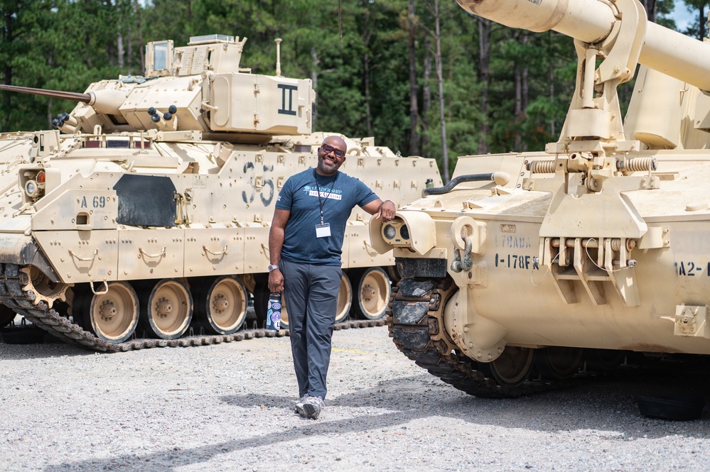 A smiling man wearing a blue t-shirt and lanyard/badge poses casually between two desert-tan military armored vehicles on a gravel surface. The vehicles appear to be Bradley Fighting Vehicles or similar tracked armored personnel carriers, marked with unit designations. The setting appears to be a military base or installation, likely during a public tour or civilian leadership program visit.