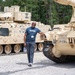 A smiling man wearing a blue t-shirt and lanyard/badge poses casually between two desert-tan military armored vehicles on a gravel surface. The vehicles appear to be Bradley Fighting Vehicles or similar tracked armored personnel carriers, marked with unit designations. The setting appears to be a military base or installation, likely during a public tour or civilian leadership program visit.