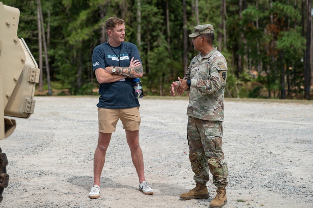 Two individuals are engaged in conversation on a gravel surface near a wooded area. One person is in civilian clothing wearing a navy blue 'Leadership South Carolina' t-shirt, while the other is in U.S. Army OCP (Operational Camouflage Pattern) uniform. A military vehicle is partially visible on the left side of the frame.