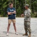 Two individuals are engaged in conversation on a gravel surface near a wooded area. One person is in civilian clothing wearing a navy blue 'Leadership South Carolina' t-shirt, while the other is in U.S. Army OCP (Operational Camouflage Pattern) uniform. A military vehicle is partially visible on the left side of the frame.