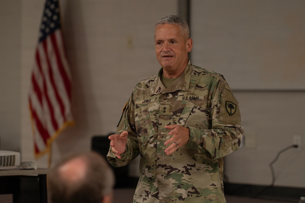 A U.S. Army Major General named Jones, identifiable by two stars on his collar and the name tape on his uniform, is addressing an audience in what appears to be a briefing or meeting room. He is wearing the Operational Camouflage Pattern (OCP) uniform with a unit patch on his left shoulder. An American flag is visible in the background.