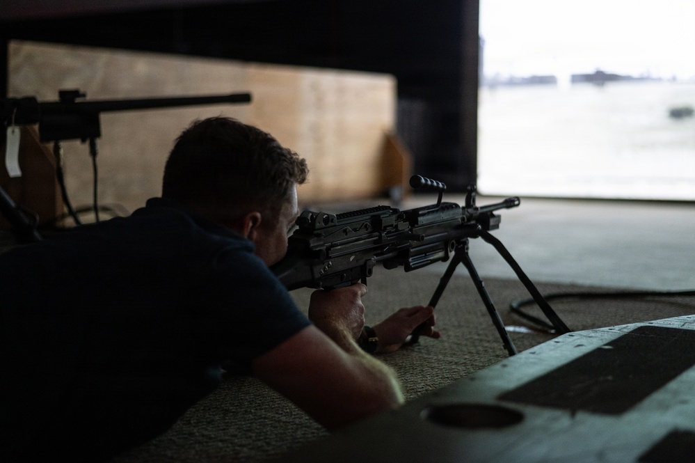 A person in a dark blue shirt is in a prone firing position at what appears to be an indoor shooting range, aiming a light machine gun mounted on a bipod. The shooter is positioned on a mat and is looking through the sights toward a brightly lit opening in the background. A second long-barreled firearm on a bipod is visible to the left.