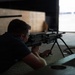A person in a dark blue shirt is in a prone firing position at what appears to be an indoor shooting range, aiming a light machine gun mounted on a bipod. The shooter is positioned on a mat and is looking through the sights toward a brightly lit opening in the background. A second long-barreled firearm on a bipod is visible to the left.