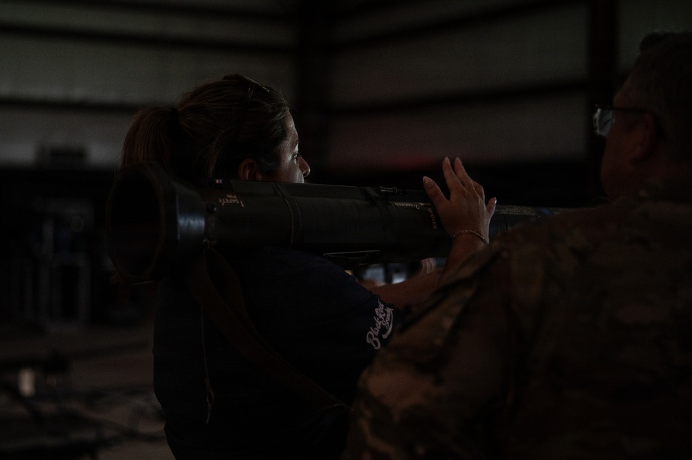 A civilian woman in a dark hoodie is being shown how to handle a military rocket launcher or similar tube-shaped weapon system by a person in military camouflage uniform. The setting appears to be an indoor facility such as a hangar or warehouse. This appears to be a demonstration or familiarization event.