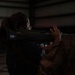 A civilian woman in a dark hoodie is being shown how to handle a military rocket launcher or similar tube-shaped weapon system by a person in military camouflage uniform. The setting appears to be an indoor facility such as a hangar or warehouse. This appears to be a demonstration or familiarization event.