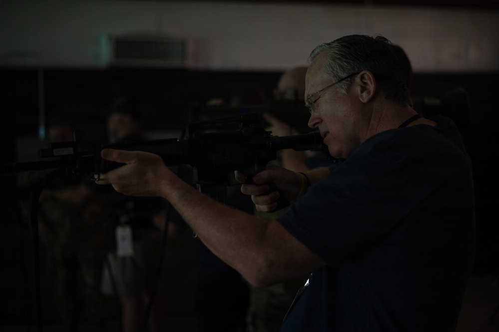 A middle-aged man with gray hair and safety glasses is aiming a rifle, likely an M4 or AR-style carbine, in what appears to be an indoor shooting range or training facility. He is wearing a dark blue shirt and is in a proper shooting stance with the weapon raised to eye level. Additional individuals are visible in the dark background, suggesting a group training or demonstration event.