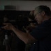 A middle-aged man with gray hair and safety glasses is aiming a rifle, likely an M4 or AR-style carbine, in what appears to be an indoor shooting range or training facility. He is wearing a dark blue shirt and is in a proper shooting stance with the weapon raised to eye level. Additional individuals are visible in the dark background, suggesting a group training or demonstration event.