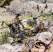 Two military personnel in different camouflage uniforms are handling a litter/stretcher on a rocky mountainside terrain. They appear to be conducting a mountain rescue or casualty evacuation training exercise, with ropes and rigging equipment visible. The setting is a rugged alpine environment with a valley and river visible in the background.