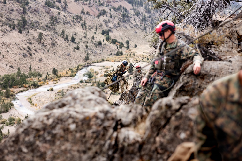Military personnel in camouflage uniforms are conducting a rappelling or rock climbing training exercise on a steep rocky cliff face. At least three individuals are visible, wearing safety helmets in red and blue, with ropes and climbing gear. The background reveals a mountainous valley with a winding road and river far below.