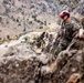 Military personnel in camouflage uniforms are conducting a rappelling or rock climbing training exercise on a steep rocky cliff face. At least three individuals are visible, wearing safety helmets in red and blue, with ropes and climbing gear. The background reveals a mountainous valley with a winding road and river far below.