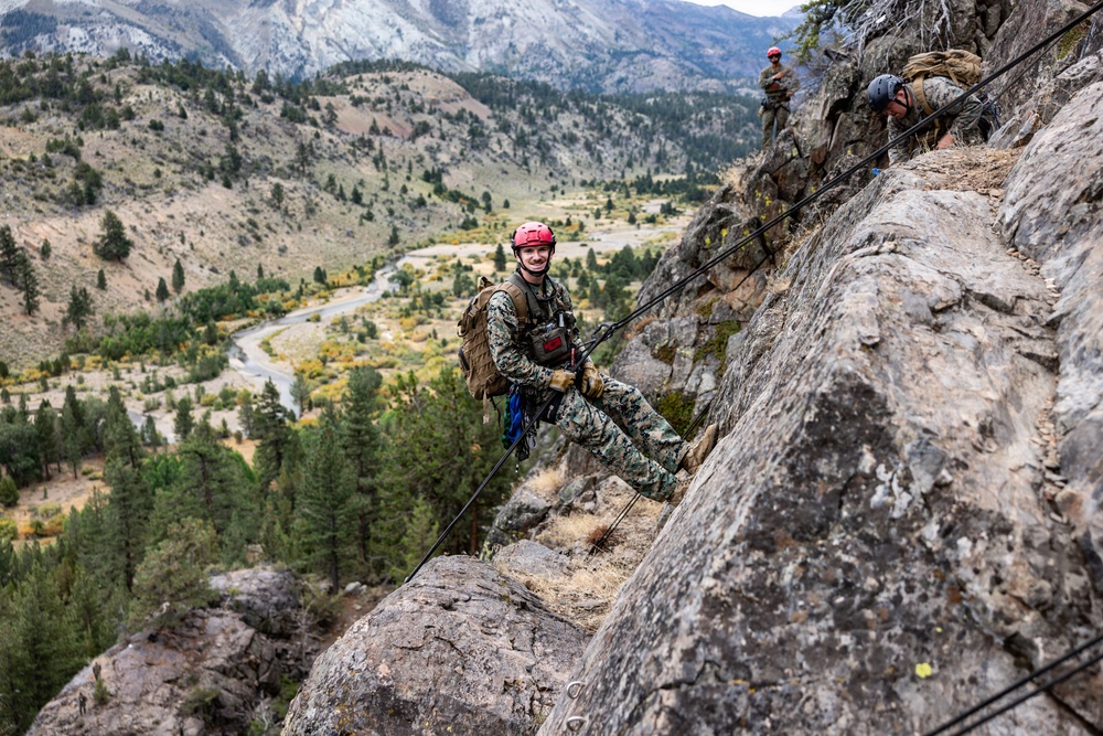 Military personnel in camouflage uniforms are conducting rappelling or rock climbing training on a steep rocky cliff face. The primary subject, wearing a red helmet and full tactical gear with a backpack, is smiling at the camera while secured by ropes on the rock face. The backdrop reveals a dramatic mountain valley with a winding road, conifer forests, and snow-capped peaks in the distance.