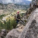 Military personnel in camouflage uniforms are conducting rappelling or rock climbing training on a steep rocky cliff face. The primary subject, wearing a red helmet and full tactical gear with a backpack, is smiling at the camera while secured by ropes on the rock face. The backdrop reveals a dramatic mountain valley with a winding road, conifer forests, and snow-capped peaks in the distance.
