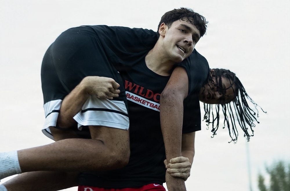 A young male athlete wearing a black 'Wildcats Basketball' t-shirt is carrying another person on his back or shoulders in what appears to be an outdoor setting. Both individuals appear to be sweaty, suggesting physical exertion during a team activity or drill. The image is captured in close-up, emphasizing the physical effort and strain on the carrier's face.