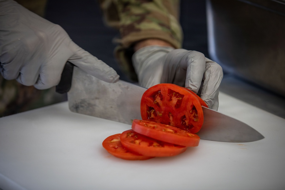 A person wearing military camouflage uniform and gray latex gloves is slicing a red tomato on a white cutting board using a large chef's knife. Several tomato slices have already been cut and are fanned out on the cutting board. The close-up shot focuses on the hands, knife, and tomato, suggesting a food preparation context in a military or institutional kitchen setting.