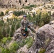 A military service member in MARPAT camouflage uniform is rappelling down a rocky cliff face using ropes and harness equipment. The scene appears to be a military training exercise conducted in a mountainous, forested environment. A valley with a winding river and conifer trees is visible in the background below.