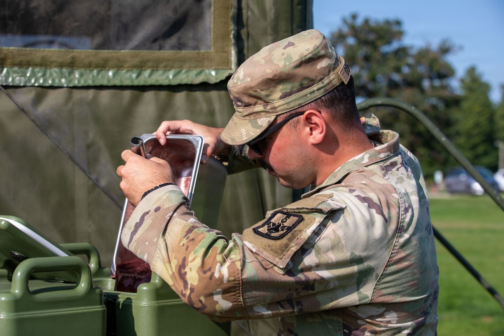 A U.S. Army soldier in Operational Camouflage Pattern (OCP) uniform is examining or pouring liquid from a metal container near a military field tent. The soldier is wearing a patrol cap and sunglasses and appears to be working with military field equipment outdoors. The setting appears to be a field training or operational environment with green military gear visible.